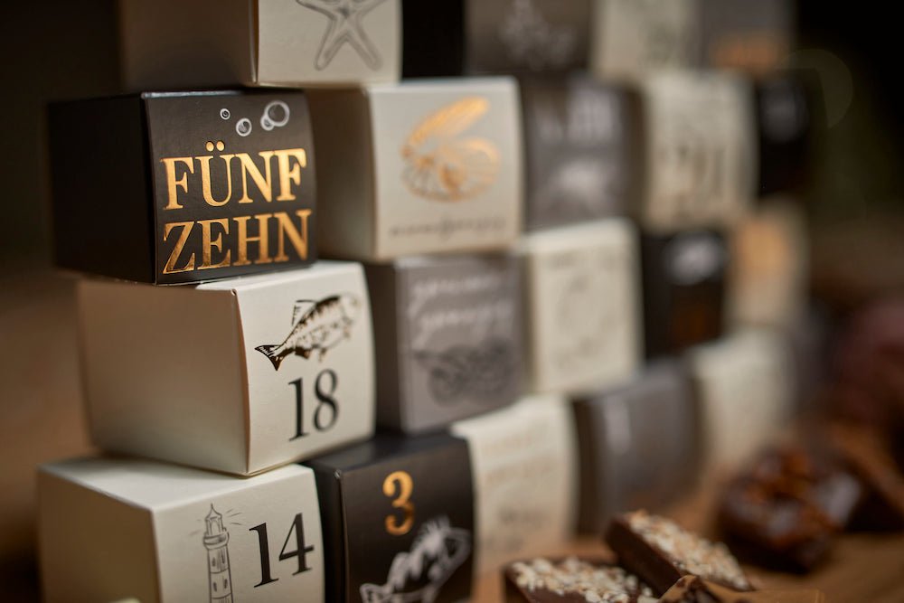 Eine Nahaufnahme gestapelter Würfel mit verschiedenen Zahlen und Symbolen, darunter das Wort „fünfzehn“, was auf Deutsch fünfzehn bedeutet, im Fokus aus dem Schokoladen-Adventskalender 12er-Set der Sylter Manufaktur Johannes King.