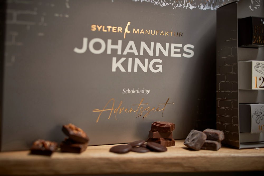 Im Vordergrund eine Reihe kunstvoll präsentierter Schokoladenstücke, im Hintergrund eine Schachtel mit der Aufschrift „Sylter Manufaktur Johannes King, Schokoladen-Adventskalender 12er Set“.