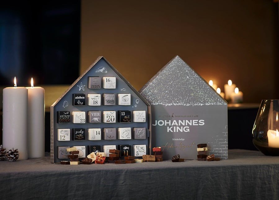 Ein festlicher Kings Schokoladen-Adventskalender 2023 in Form von Häusern mit offenen Schubladen, die den Blick auf Edelschokoladen freigeben, flankiert von brennenden Kerzen und einer Laterne.
