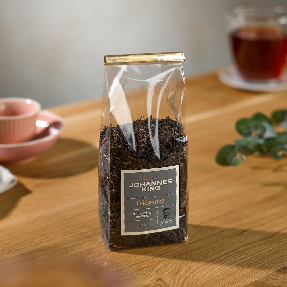 Eine durchsichtige Plastikverpackung mit losem Blattwerk „Kings Friesenteemischung vom Söl’ring Hof“, auf einem Holztisch platziert, im Hintergrund eine Tasse Tee und eine Untertasse.
