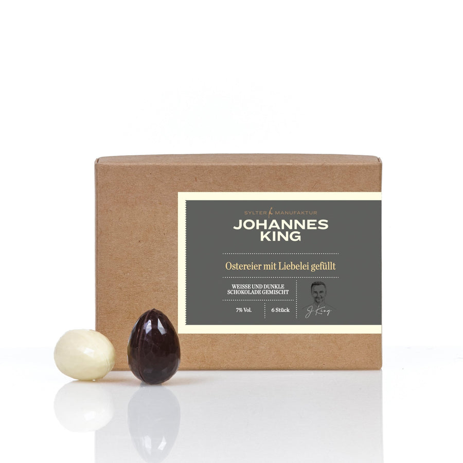 Eine verpackte Schachtel mit der Aufschrift „Sylter Manufaktur“ enthält sechs mit Ganache gefüllte Schokoladen-Ostereier, ausgestellt neben zwei Schokoladeneiern, einem weißen und einem dunklen, vor einem weißen Hintergrund.