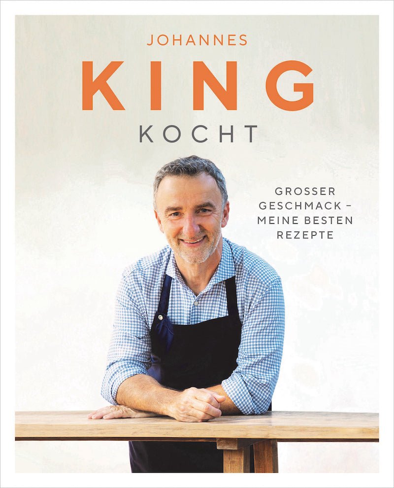 Ein lächelnder Gourmetkoch, Johannes King, mit blauer Schürze steht hinter einer Holztheke auf dem Cover eines Kochbuchs mit dem Titel „Sylter Manufaktur kocht – Kochbuch“, was übersetzt „Johannes kocht“ bedeutet.