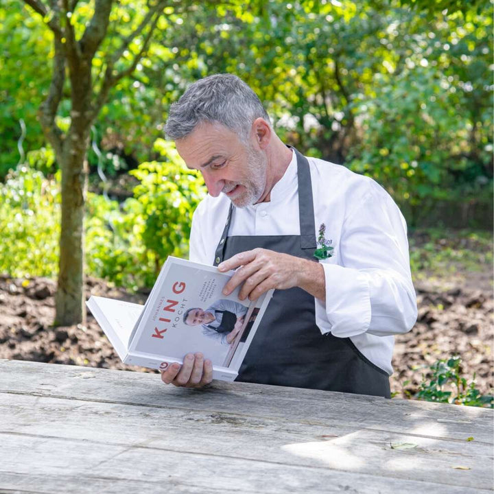 Ein König Kocht trägt einen weißen Mantel mit Schürze und lächelt, während er an einem Holztisch im Freien, umgeben von viel Grün, ein Buch liest.