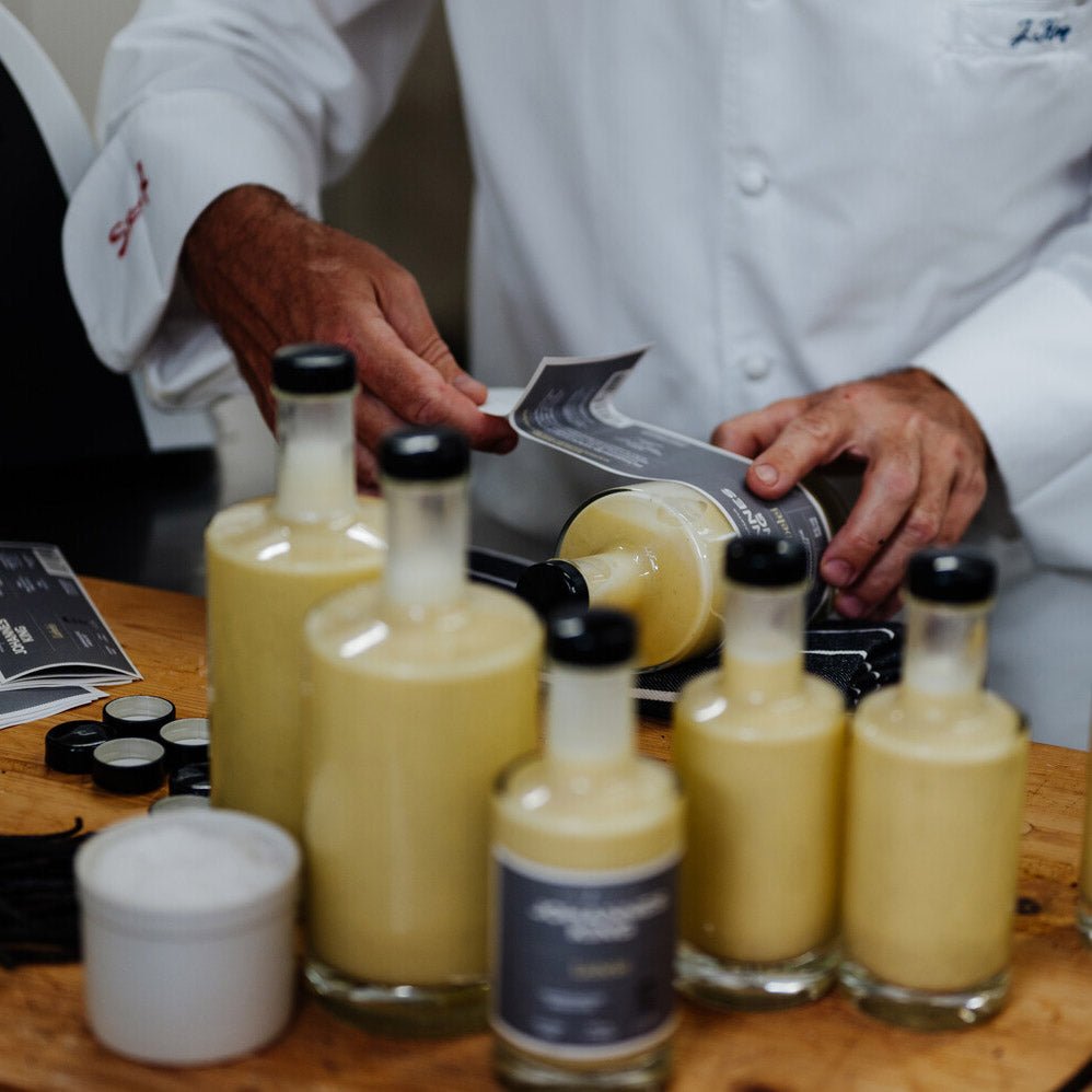 Ein Koch gießt Sylter Manufaktur Johannes King Liebelei-Duo, eine gelbe Flüssigkeit, in Glasflaschen, die Teil einer Reihe ähnlich gefüllter Flaschen auf einer Küchentheke sind.