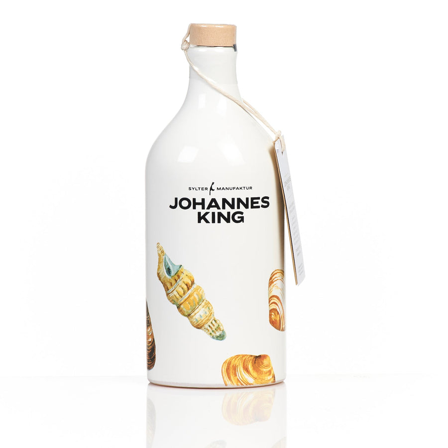 Eine weiße, undurchsichtige Flasche mit „Johannes King“-Etikett, verziert mit Bildern von Gebäck und Olivenöl extra vergine in der Tonkrug Muschel-Edition, mit Holzstopfen und einer Sylter Manufaktur.