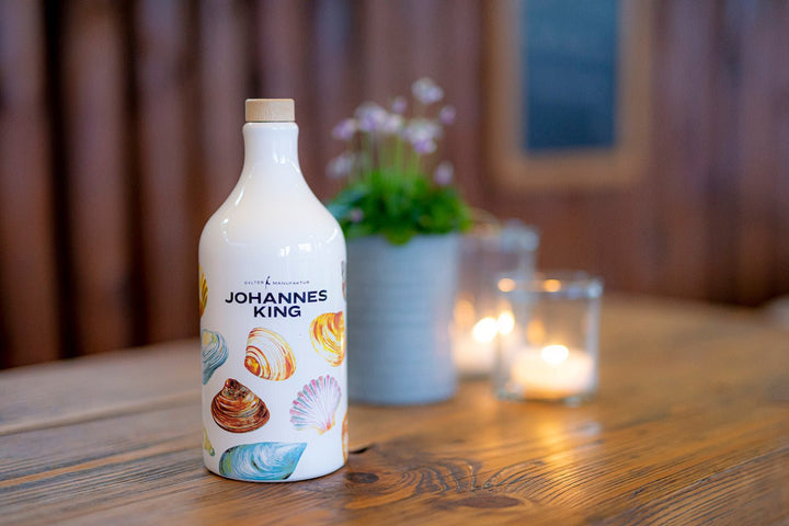 Eine Flasche Sylter Manufaktur Olivenöl extra vergine im Tonkrug *Muschel-Edition* auf einem Holztisch, flankiert von einer Topfpflanze und zwei brennenden Kerzen im Hintergrund.