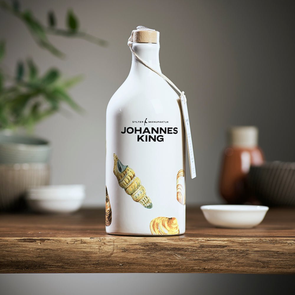 Eine weiße Flasche mit Sylter Manufaktur-Etikett, verziert mit Muschelbildern, auf einer Holzoberfläche platziert. Am Flaschenhals hängt ein Etikett.