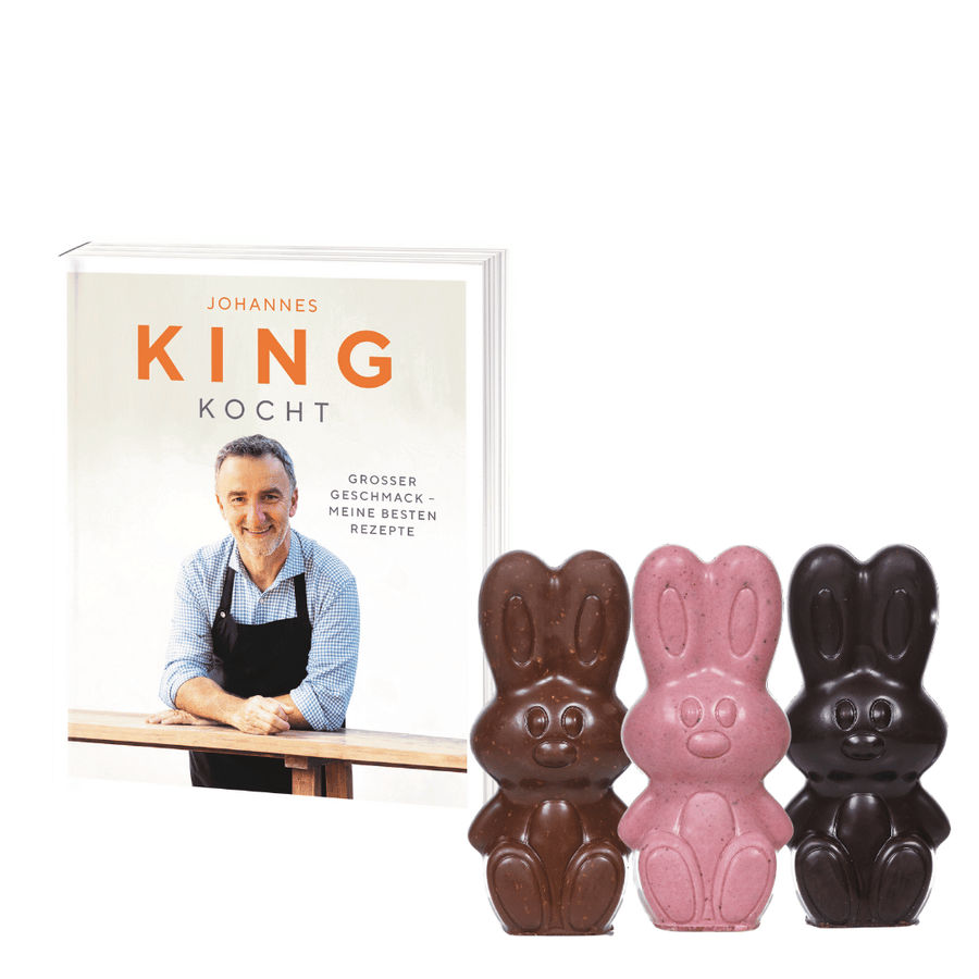 Ein Oster-Kochbuch-Set der Sylter Manufaktur mit dem Titel „Johannes King Kocht“ wird neben drei Schokohasen gezeigt, einer in weißer Schokolade, einer in Milchschokolade und ein dritter in dunkler Schokolade.