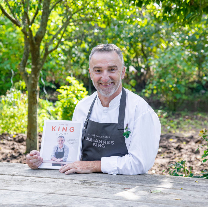 Ein lächelnder Koch mit Schürze hält ein „Für Gourmet-Mamis“ mit seinem Porträt auf dem Cover in der Hand und posiert im Freien mit viel Grün im Hintergrund.