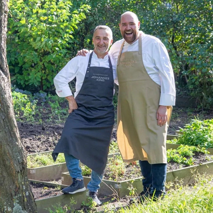 Zwei Köche, einer mit einer weißen Profi-Kochschürze der Sylter Manufaktur Johannes King und der andere mit einer hellbraunen Schürze der Sylter Manufaktur, stehen lächelnd in einem Garten.