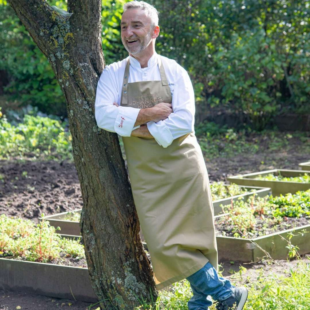 Ein Chef, der eine Profi-Kochschürze *Sylter Manufaktur* beige trägt, steht in einem Garten mit überkreuzten Armen, lehnt an einem Baum und lächelt. Sylter Manufaktur Johannes König.
