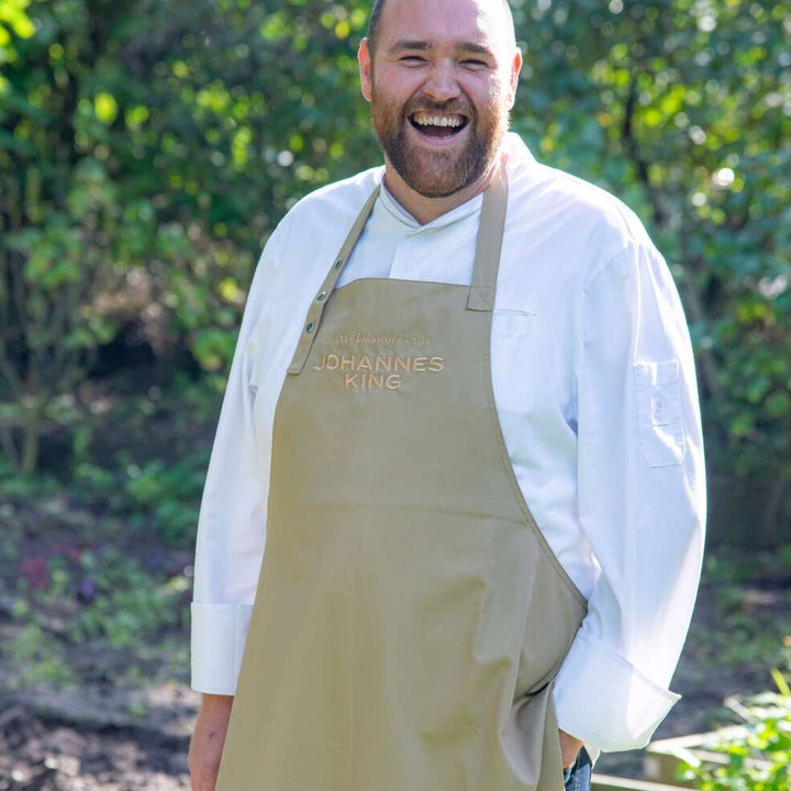 Ein fröhlicher Koch im weißen Kittel und mit einer personalisierten Schürze der Sylter Manufaktur Johannes King steht draußen und lacht.
