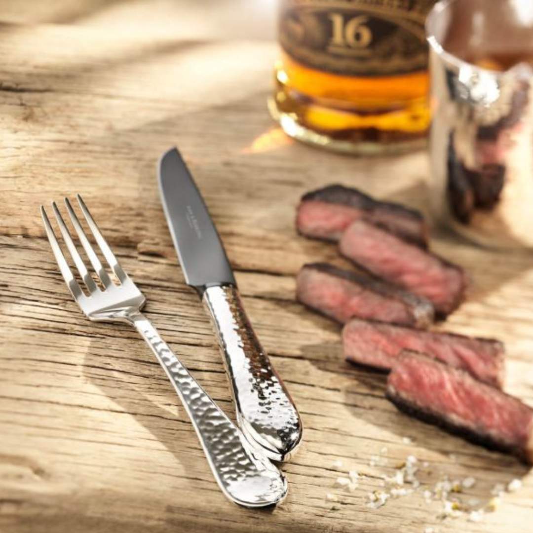 Ein Messer und eine Gabel aus dem Robbe & Berking Grillbesteck 8-tlg. Die Martelé-Kollektion ruht auf einer Holzoberfläche neben geschnittenen Steakstücken, im Hintergrund ein Glas Whiskey.