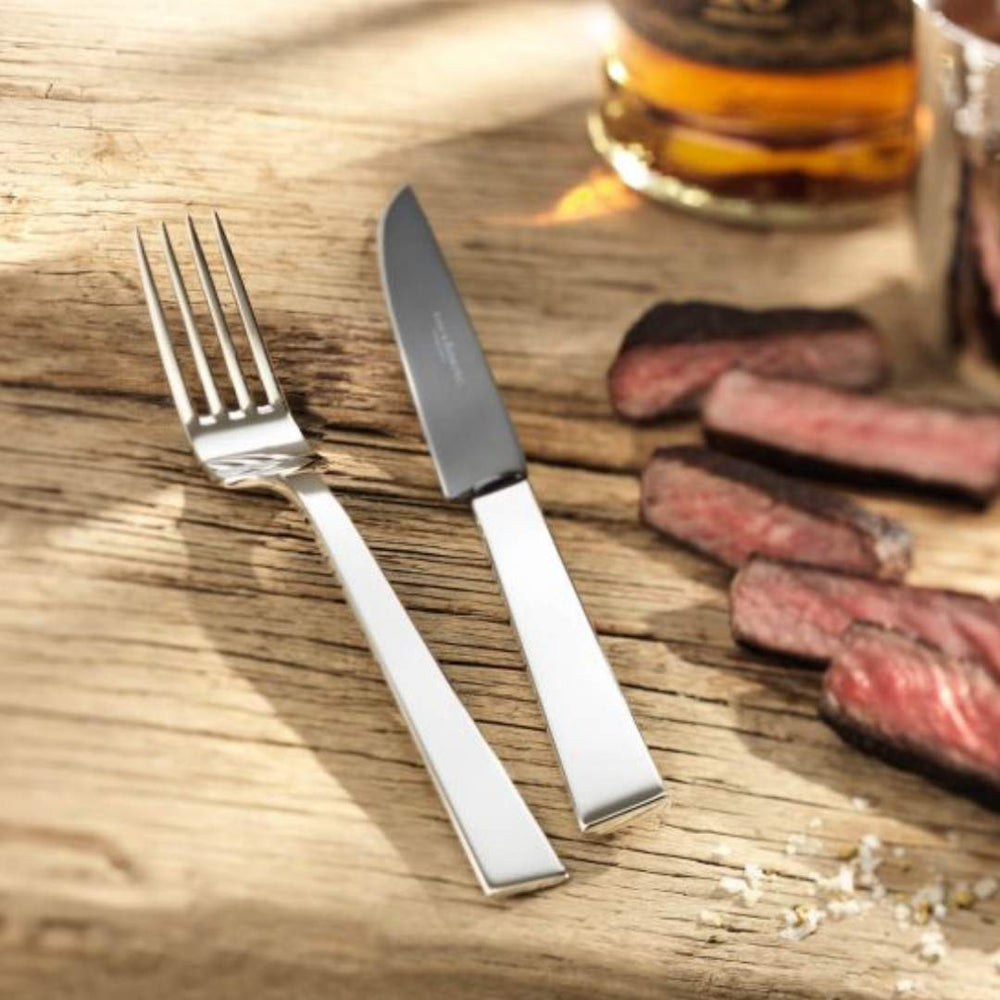 Ein Robbe & Berking Grillbesteck 8-tlg. Riva-Steakmesser und -gabel auf einem Holztisch neben gekochten Steakscheiben und einem Glas Whiskey.