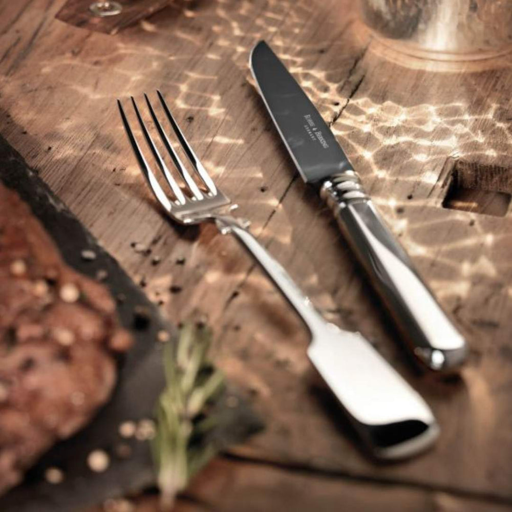 Ein Robbe & Berking Grillbesteck 8-tlg. Alt-Spaten-Gabel und -Messer mit Metallgriffen liegen nebeneinander auf einer Holzoberfläche, im Hintergrund ein verschwommenes Steak, das an eine Mahlzeit denken lässt.