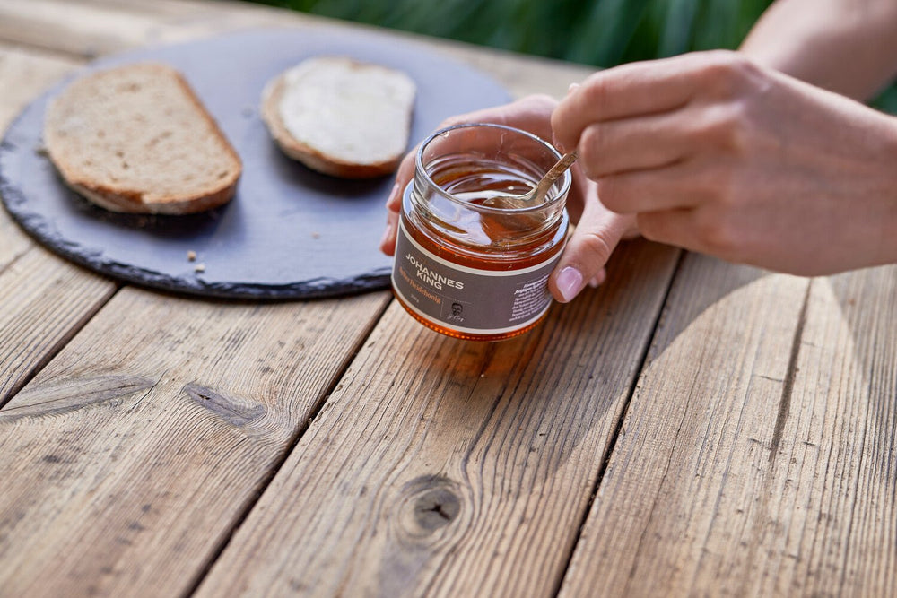 Eine Person schöpft Sylter Heidehonig aus einem Glas mit einem hölzernen Honiglöffel auf eine Brotscheibe auf einem Holztisch.