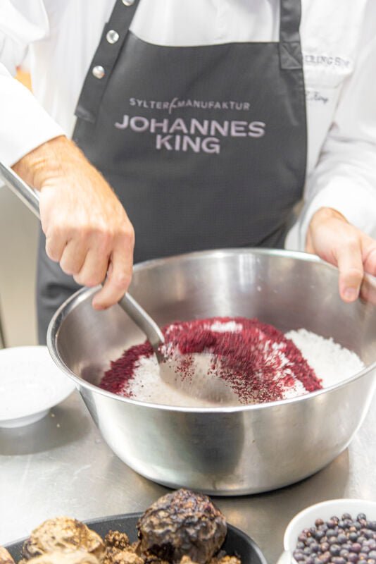Ein Koch mit einer personalisierten Schürze mischt Zutaten, darunter Sylter Rote Bete-Salz, in einer großen Metallschüssel, wahrscheinlich während eines Koch- oder Backvorgangs.
