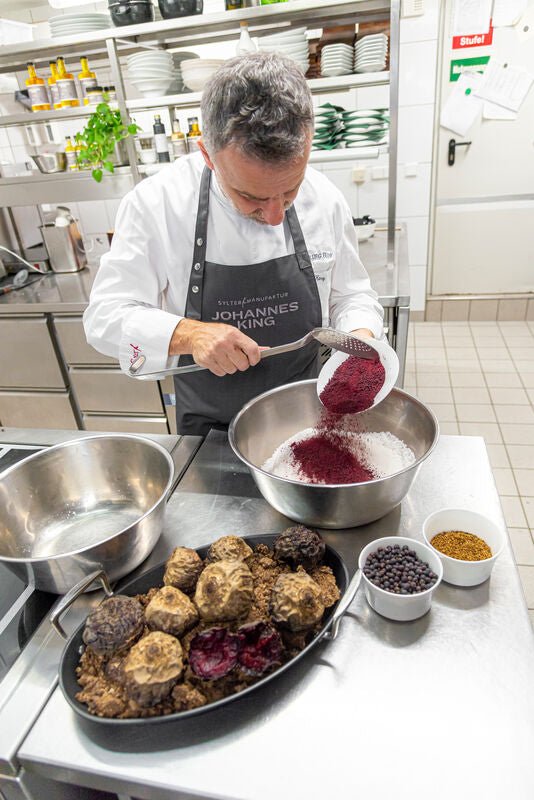 Ein Koch siebt in einer Profiküche eine violette Substanz in eine Metallschüssel, neben ihm steht auf der Arbeitsplatte ein Topf mit Trüffeln, angereichert mit Sylter Manufaktur Rotes Bete-Salz.