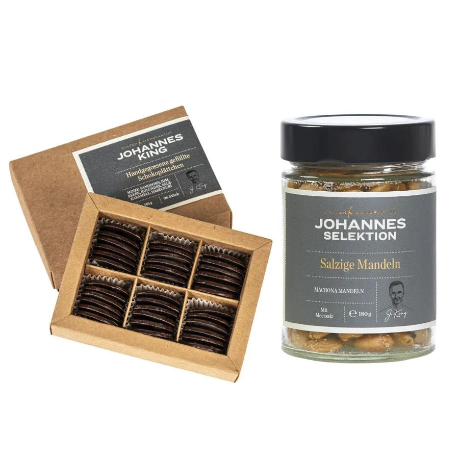Ein Set mit Gourmet-Produkten, darunter eine Schachtel Leckere Snacks aus der Manufaktur mit Schokoladenüberzug und der Aufschrift „Johannes König“ sowie ein Glas „Salzige Mandeln“ aus der Sylter Manufaktur.