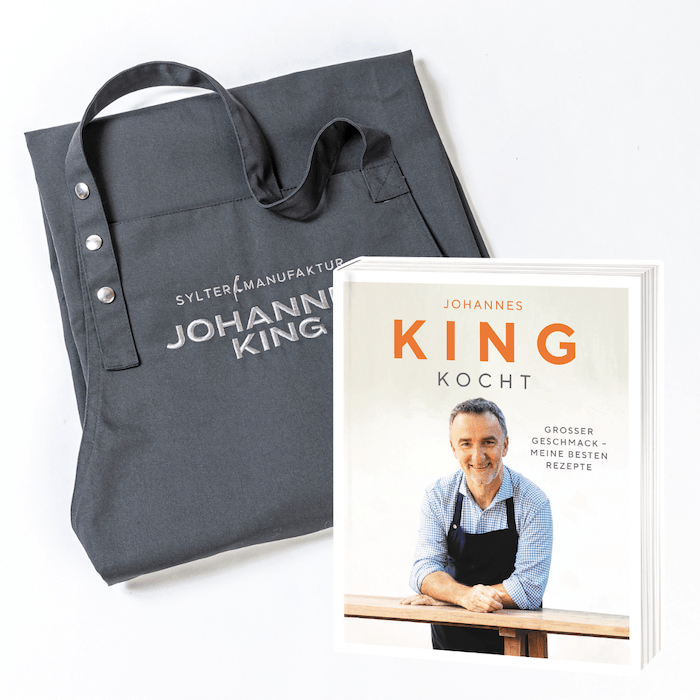 Ein dunkles Geschenk-Set für Kochfans, bestehend aus einer Tasche mit der Aufschrift „Sylter Manufaktur“ neben einem Kochbuch mit dem Titel „King Kocht“.