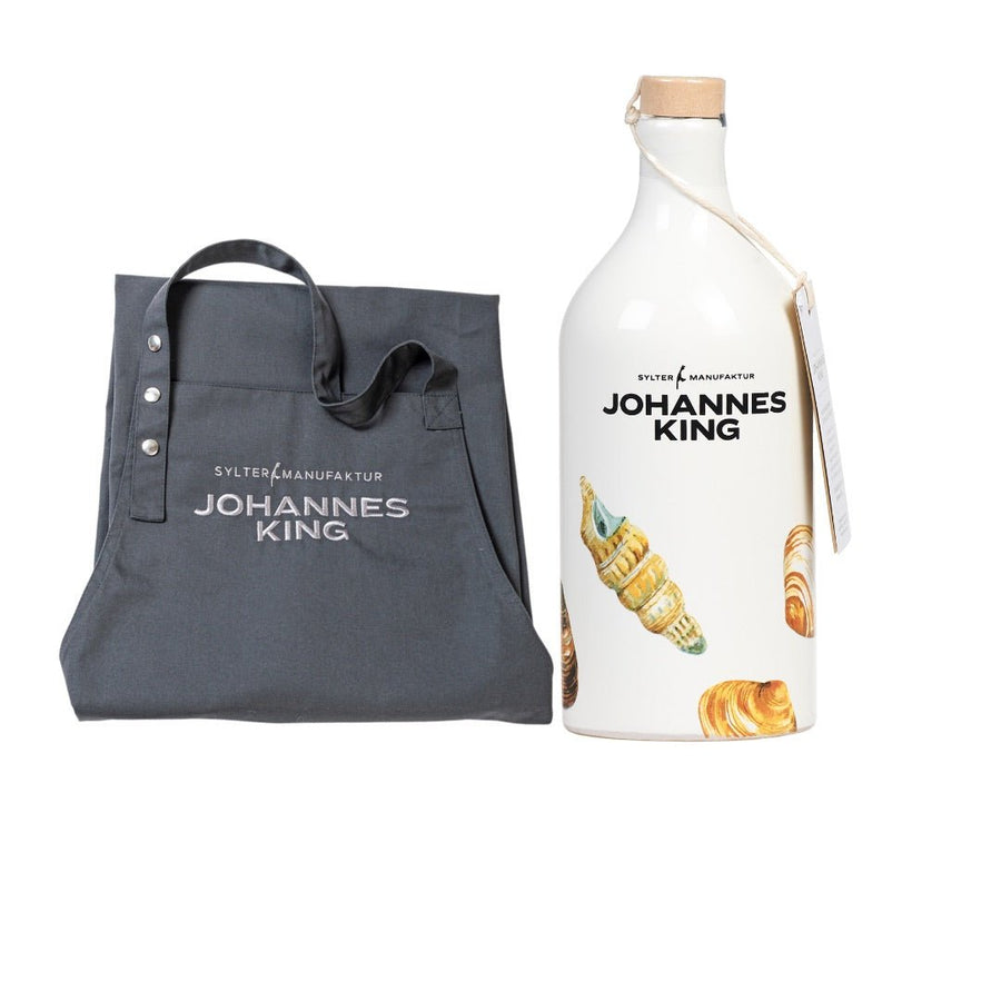 Eine graue Einkaufstasche mit „Sylter Manufaktur“-Schriftzug neben einer weißen Flasche Olivenöl mit „Sylter Manufaktur“-Schriftzug und Bildern von Gebäck.