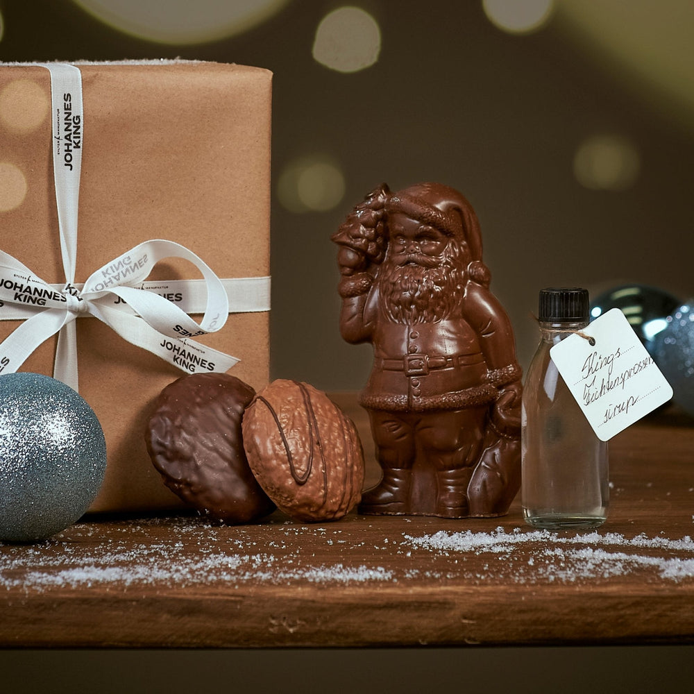 Ein festliches Arrangement mit einem Weihnachtlichen Schokotraum der Sylter Manufaktur, einer Geschenkbox mit weißem Band, glitzernden Kugeln, einer Walnuss, Nürnberger Elisen-Lebkuchen und einem.