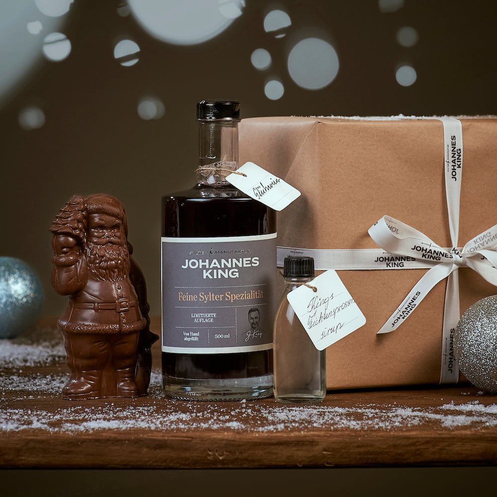 Eine Flasche Sylter Manufaktur Glühwein Geschenkset neben einem Schokoladen Geschenk, einem verpackten Geschenk und Weihnachtsdekorationen auf einer Holzoberfläche mit einer Prise Puderzucker.