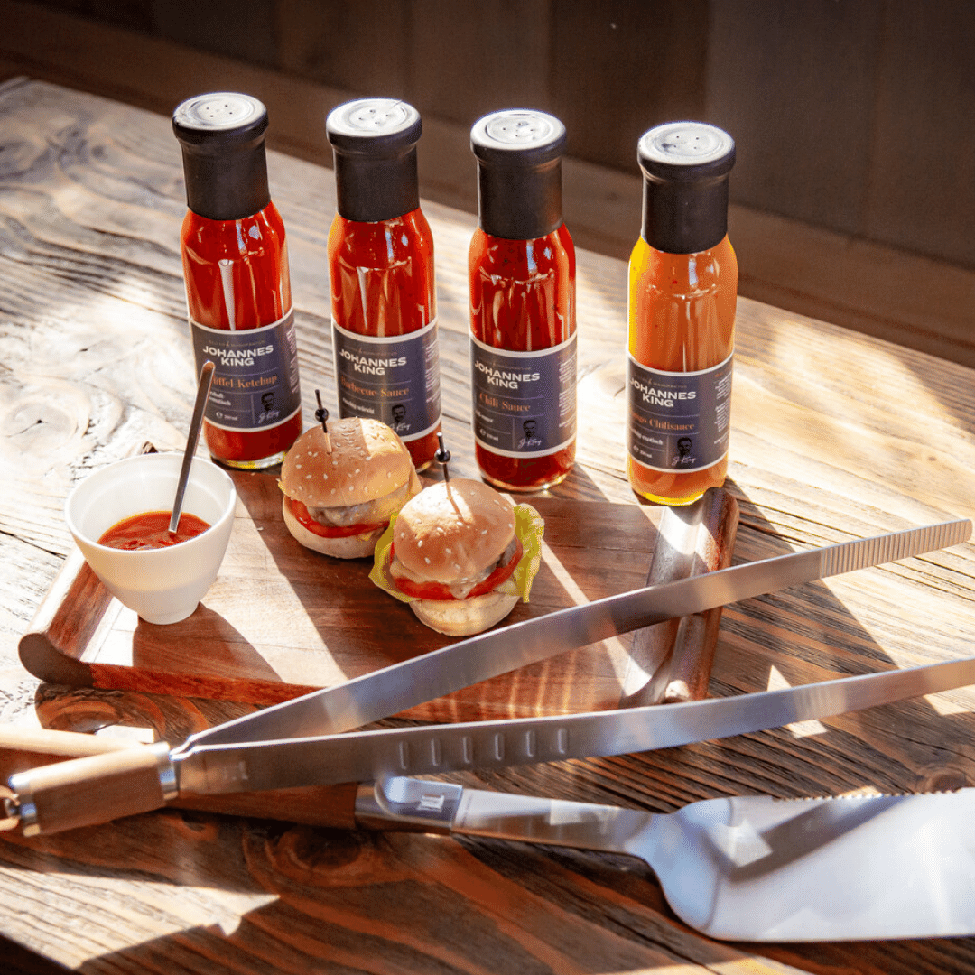 Vier Flaschen verschiedener Saucen mit dem Markennamen „Johannes“ auf einem Holztisch, dazu ein kleiner Hamburger, eine Schale Sylter Manufaktur Trüffel-Ketchup und ein Küchenset.