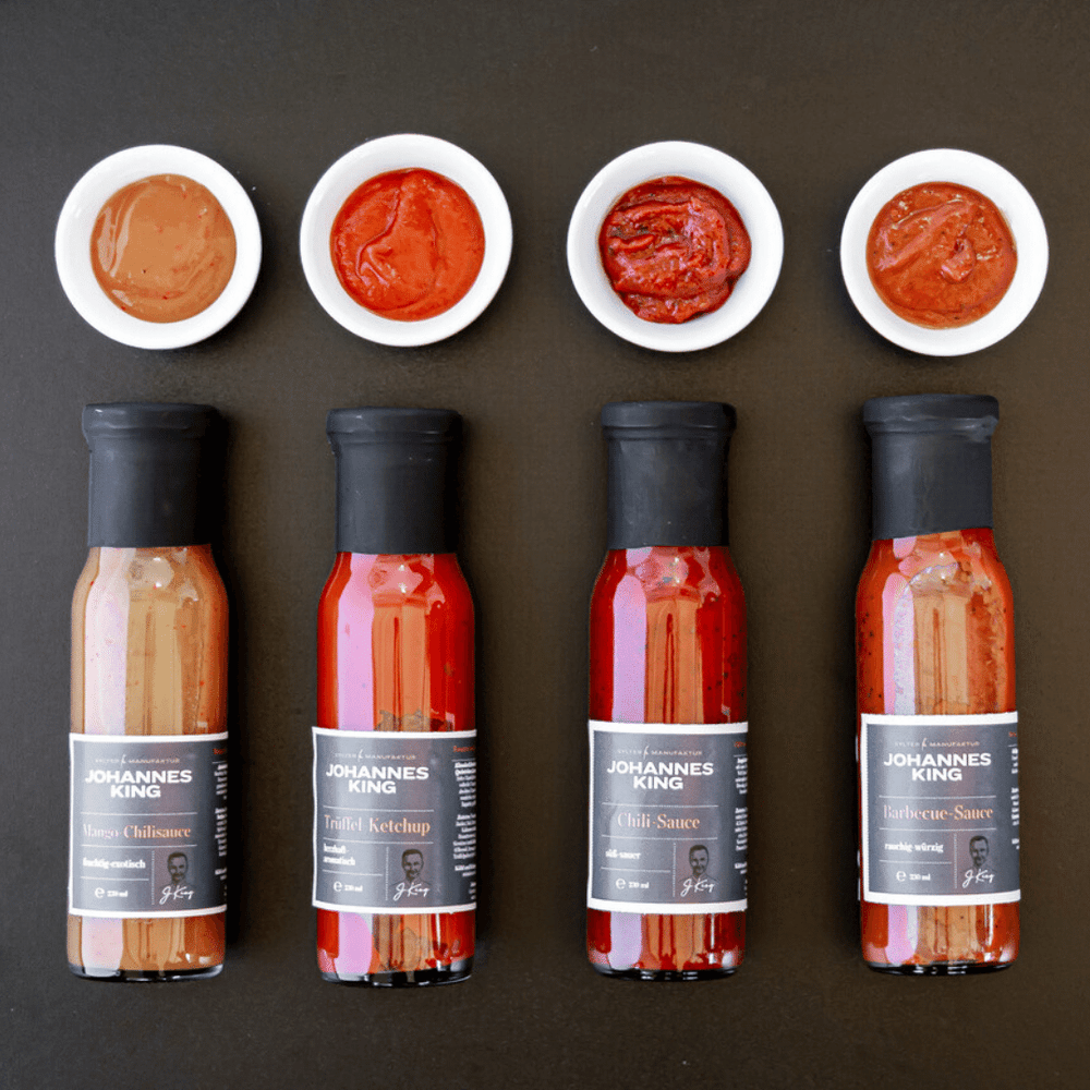 Vier Flaschen Sylter Manufaktur süße Chili-Sauce horizontal ausgerichtet mit entsprechenden Saucenproben, darunter süße Chili-Sauce, darüber jeweils auf weißen Untertassen platziert, präsentiert auf einem dunklen Hintergrund, perfekt für Fusion-Küche.