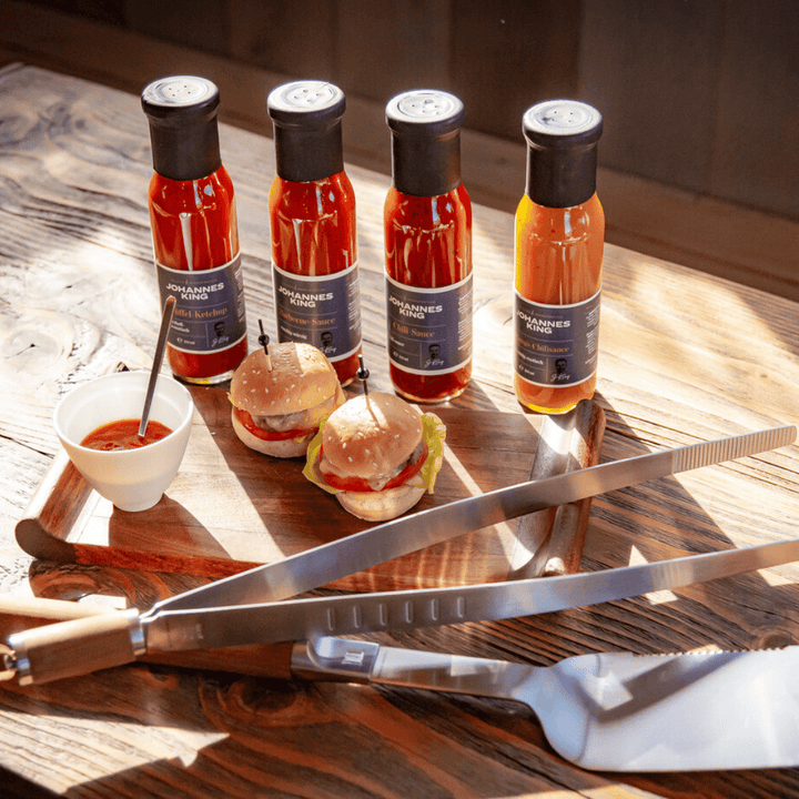 Vier Flaschen Sylter Manufaktur Mango Chili-Sauce mit der Aufschrift "Johannes" stehen auf einem Holztisch neben einem kleinen Hamburger und einer Schale mit Sauce. Zwei große Küchenmesser liegen gekreuzt in