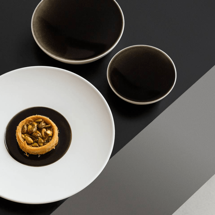 Eine minimalistische Präsentation von Speisen mit einem einzelnen Baklava-Stück in der Mitte einer weißen Hering Berlin Schale mit außenliegender freimodellierter Struktur Evolution – Obsidian, flankiert von zwei dunklen Schalen auf einem zweifarbigen Hintergrund.