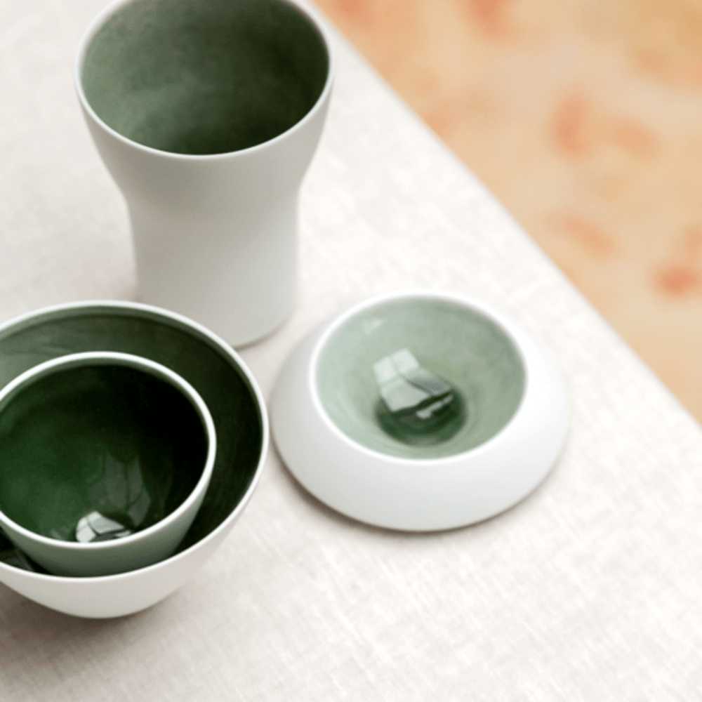 Ein Satz Hering Berlin Schale mit außenliegender freimodellierter Struktur Evolution – Smaragdgrünes Geschirr mit glänzender, tiefgrüner Glasur-Oberfläche, ordentlich angeordnet auf einer beigen strukturierten Oberfläche.