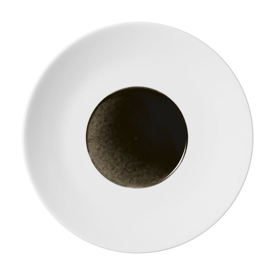 Ein minimalistisches Bild mit einer kleinen Menge dunkler Flüssigkeit, wahrscheinlich Sojasauce, in der Mitte eines groben Obsidian-Tellers von Hering Berlin Coupeteller.