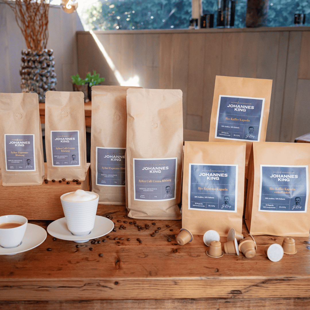 Verschiedene Kaffeeprodukte der Sylter Manufaktur, darunter abgepackte Kaffeebohnen und kompostierbare Bio Espresso Kapseln, präsentiert mit einer Espressotasse auf einem Holztisch.