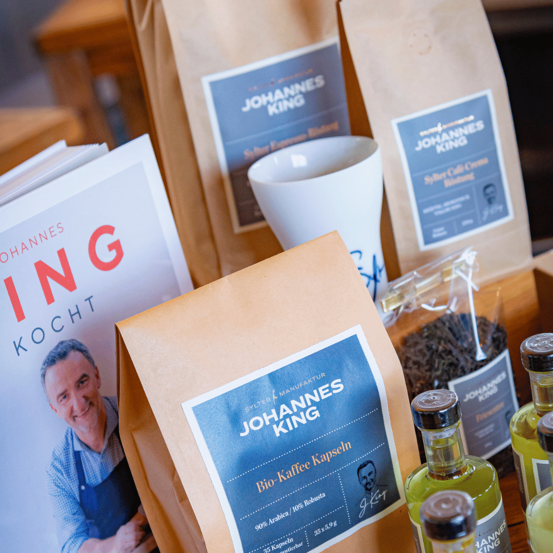 Verpackte Gourmet-Lebensmittel, darunter Bio-Kaffeekapseln der Sylter Manufaktur, die mit Nespresso-Systemen kompatibel sind, und Soßenflaschen, mit einem Branding, das das Bild und den Namen eines Mannes auf einem Holzschild zeigt.