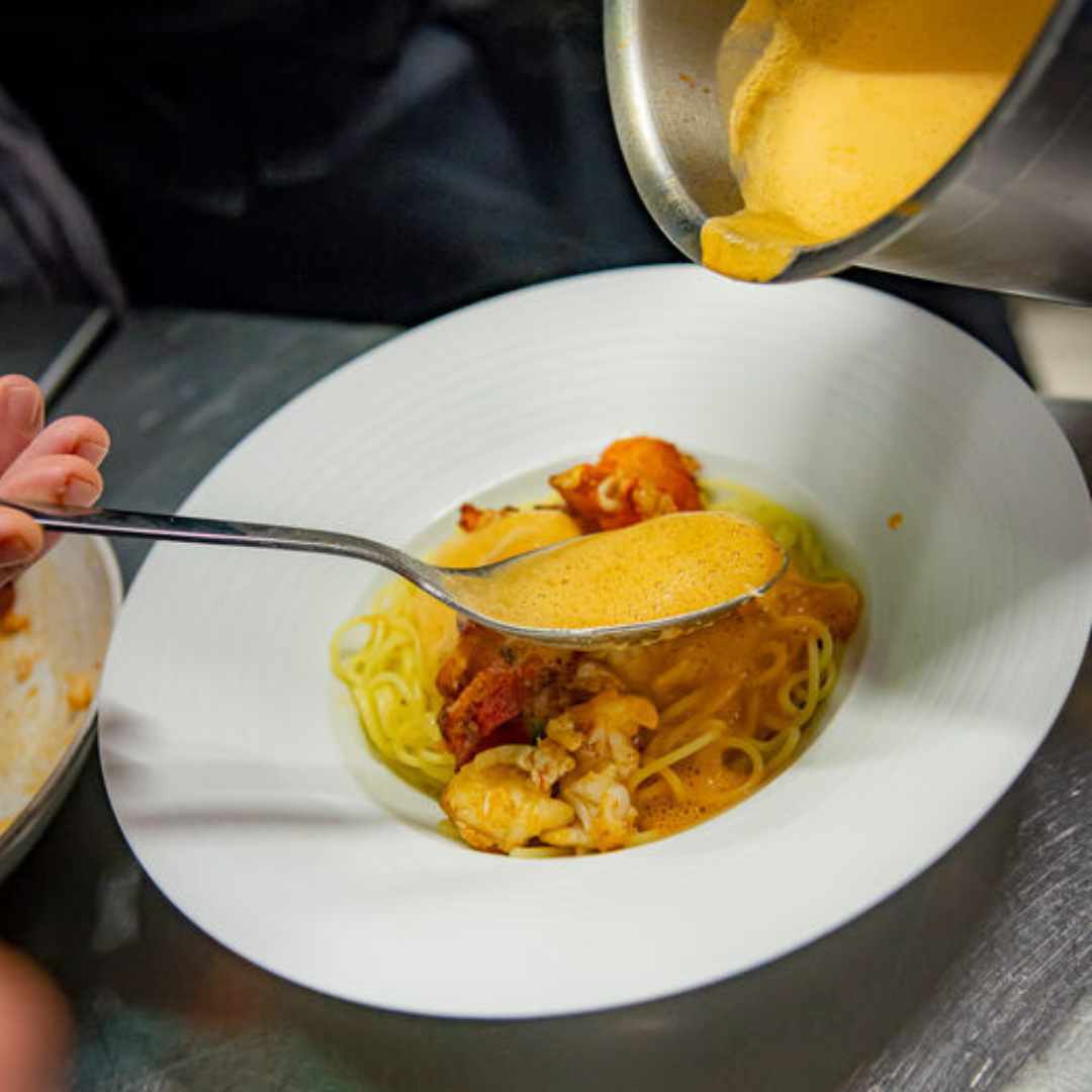 Koch übergießt Sylter Pasta-Set *Hummer* und Garnelengericht in einer weißen Schüssel der Sylter Manufaktur mit Sauce.