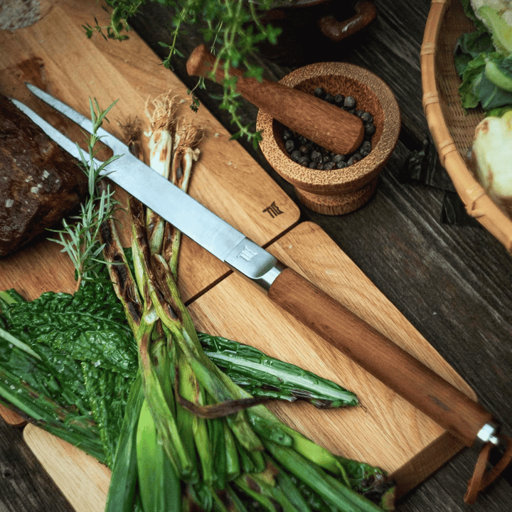 Eine Fiskars Norden Grillgabel mit frischem grünem Gemüse und einem Kochmesser sowie Mörser und Stößel mit Pfefferkörnern.