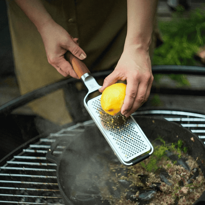 Eine Person reibt eine Zitrone über einem Barbecue-Grill, während im Hintergrund Rauch aufsteigt, mit einer Fiskars Norden Handreibe fein von Fiskars Group.