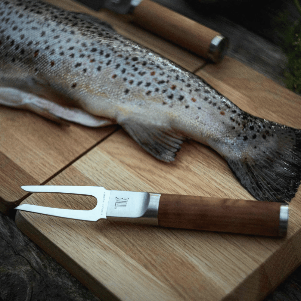 Eine frisch gefangene Forelle liegt neben einer Fiskars Norden Fischgabel auf einer Holzoberfläche.