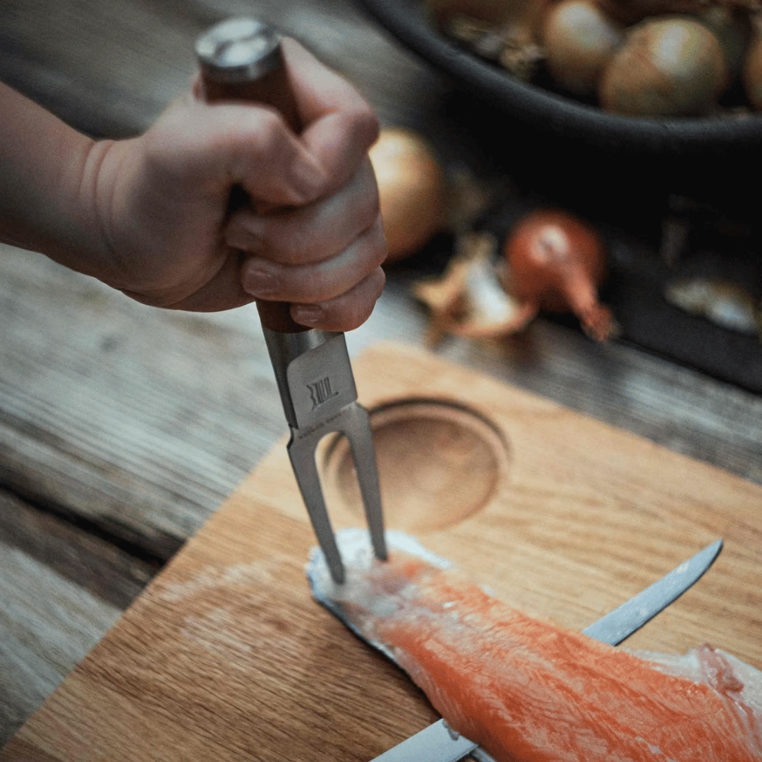 Eine Person schält mit einer Fiskars Norden Fischgabel eine Karotte über einem hölzernen Schneidebrett, im Hintergrund sind Zwiebeln in einer Schüssel.