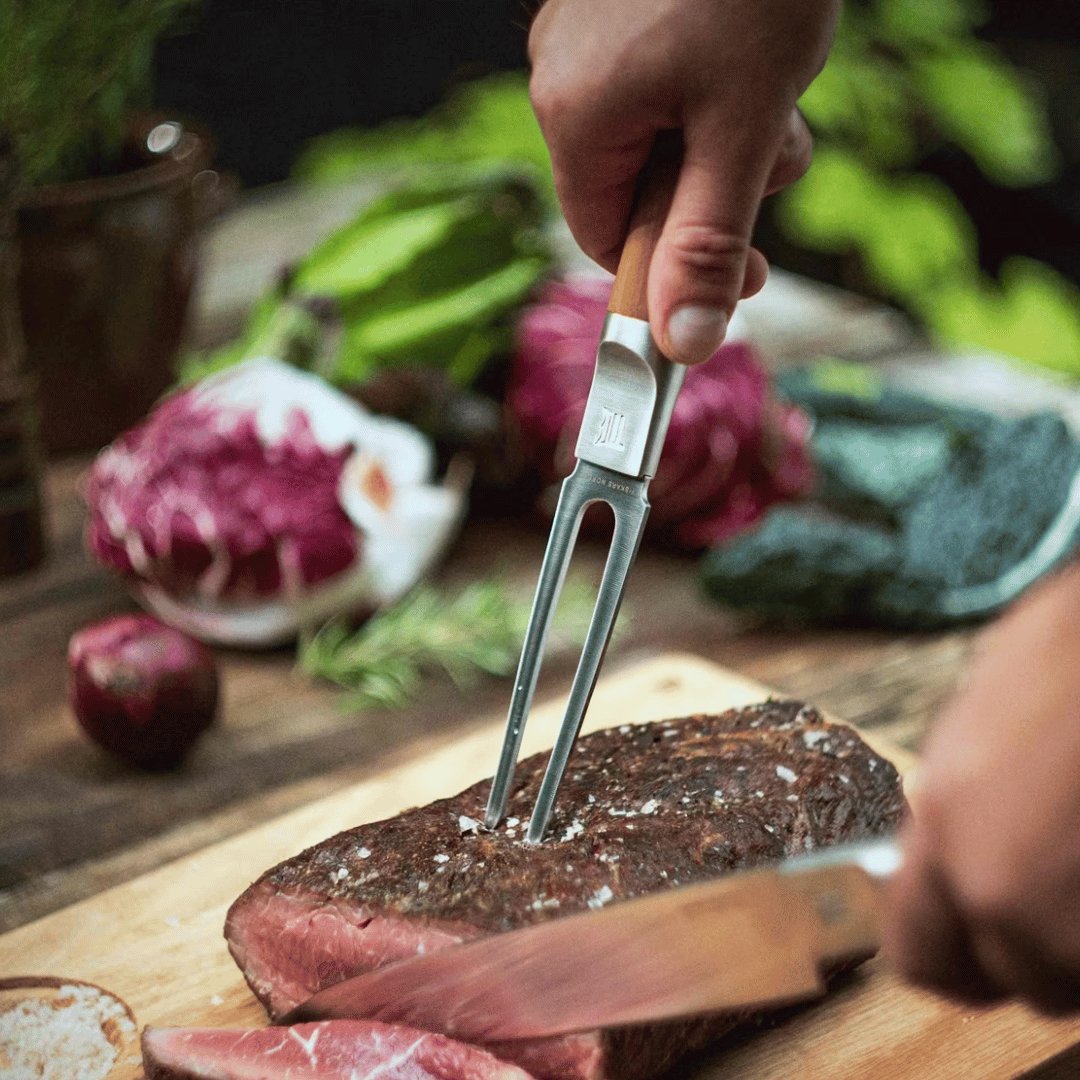 Eine Person schneidet mit einer Fiskars Norden Tranchiergabel und einem Messer ein gekochtes Steak auf einem Holzbrett, umgeben von verschiedenen Gemüsesorten.