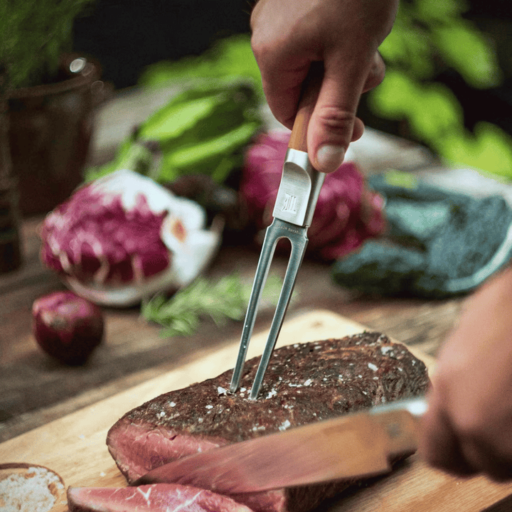 Eine Person schneidet mit einer Fiskars Norden Tranchiergabel und einem Messer ein gekochtes Steak auf einem Holzbrett, umgeben von verschiedenen Gemüsesorten.