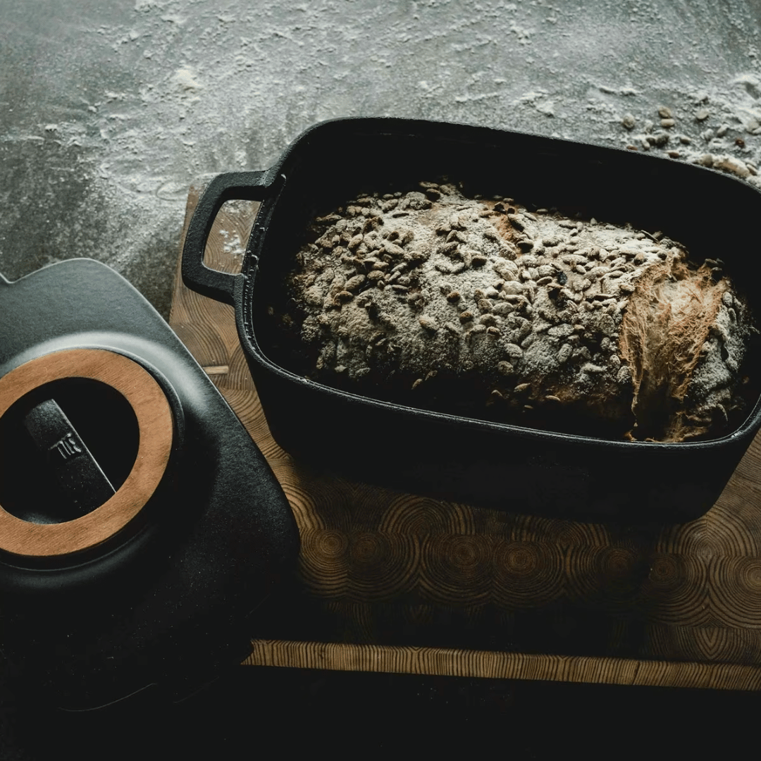 Ein frisch gebackener Laib Brot liegt in einem ovalen Fiskars Norden Topf (5,0 l) aus Gusseisen, der auf einem mit Mehl bestäubten Holzbrett steht.