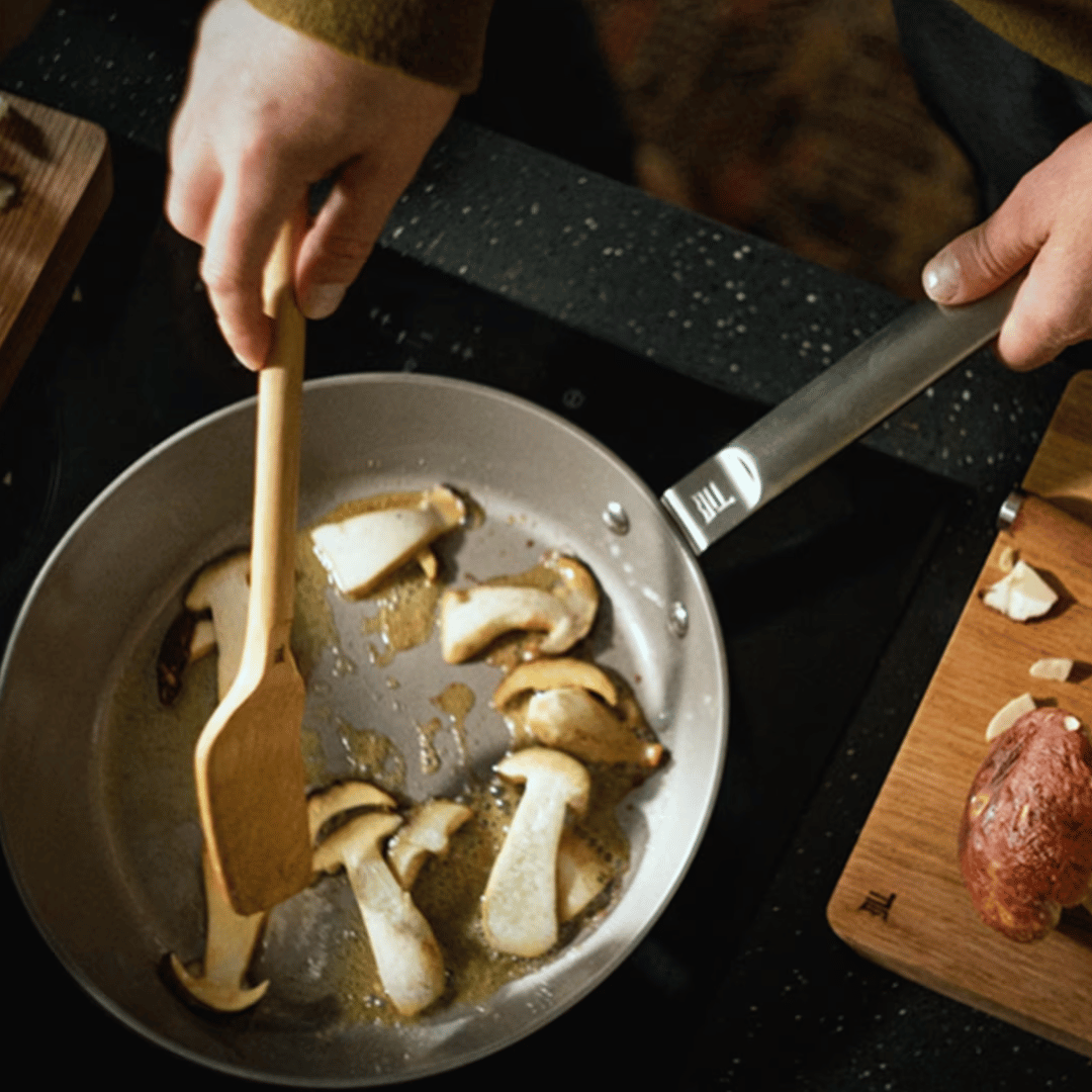 Eine Person brät geschnittene Champignons in einer beschichteten Fiskars Norden Steel Bratpfanne (28 cm) mit einem Holzspatel an. Daneben liegt ein Schneidebrett mit einer Kartoffelscheibe.