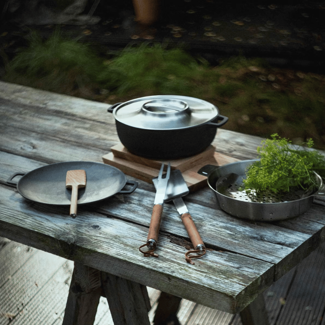 Eine rustikale Outdoor-Kochszene mit einem gusseisernen Topf auf einem Holztisch, dazu ein Teller, Utensilien und eine Schüssel mit Kräutern und Sylter Manufaktur Mango-Chili-Sauce.
