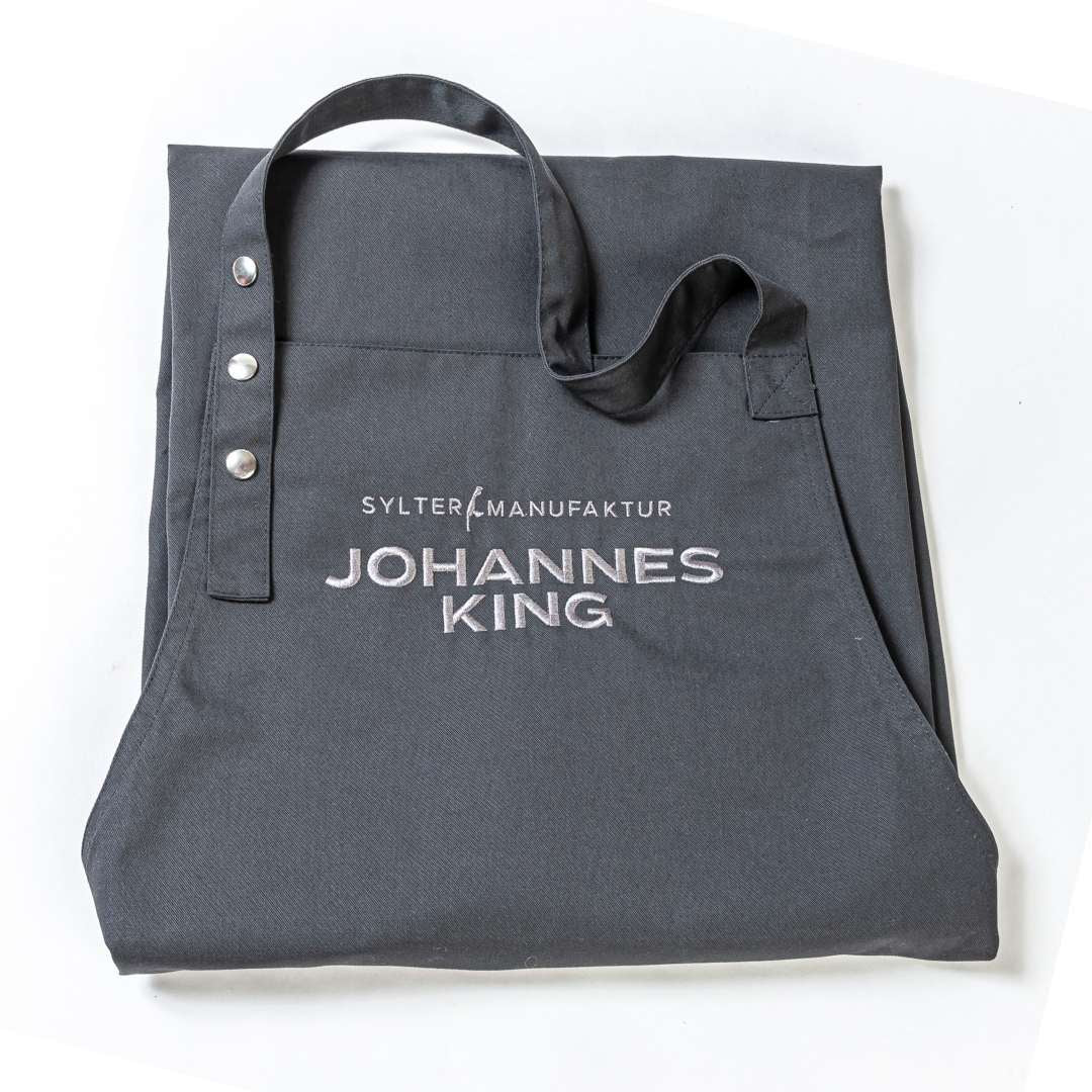 Eine schwarze Tragetasche mit dem Aufdruck „Sylter Manufaktur Johannes King – Geschenk für Grillmeister“ auf der Vorderseite vor weißem Hintergrund.
