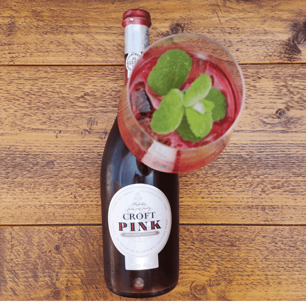 Eine Flasche Croft Pink Portwein liegt auf einer Holzoberfläche, darauf ein Glas Croft Rosé-Portwein mit Stiel, garniert mit Minzblättern.