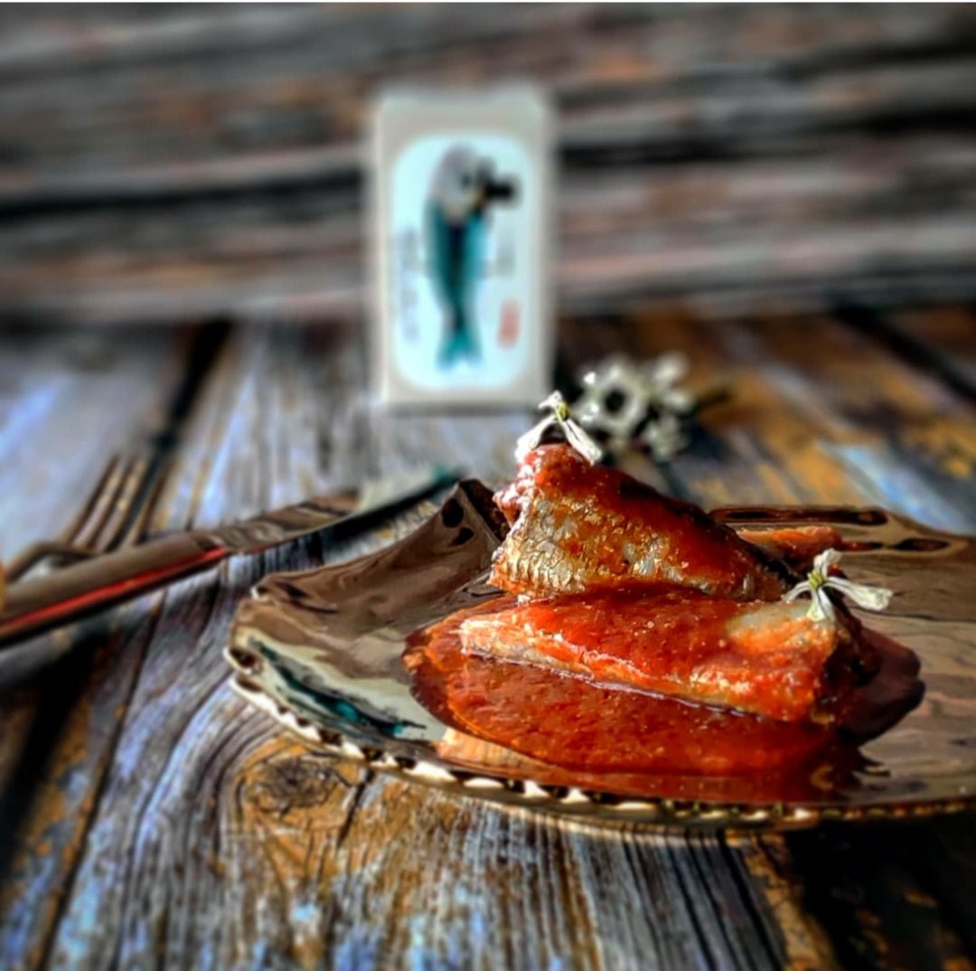 Eine plattierte Scheibe gegrillten Lachs, serviert mit Kräutern, mit einer Spielkarte im Hintergrund auf einer rustikalen Holztischplatte und Jose Gourmet Sardinen in Tomatensoße am Rand.