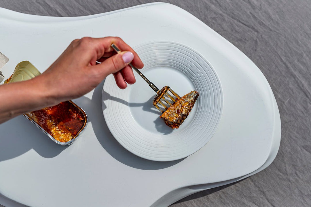Die Hand einer Person platziert mit einer Gabel eine Jose Gourmet Sardinen in Tomatensoße auf einem weißen Teller aus einer offenen Dose hochwertiger Fischdosen daneben.