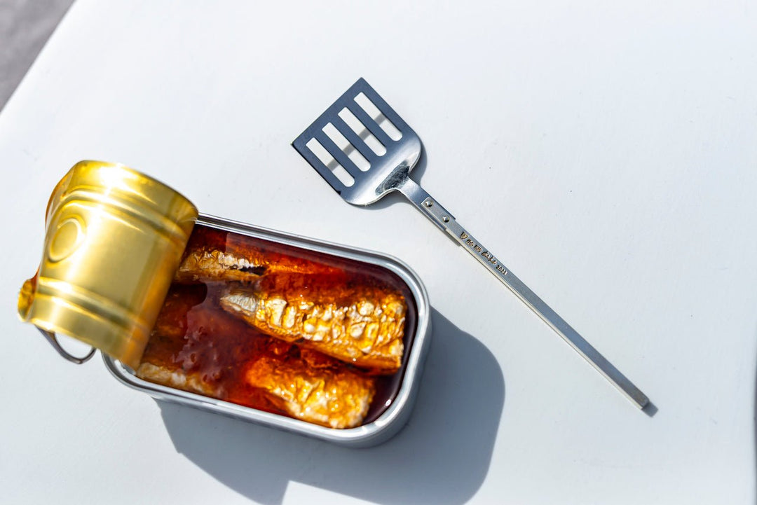Eine geöffnete Dose Jose Gourmet Sardinen in Tomatensauce, daneben liegt ein Metallspatel auf einer weißen Fläche.